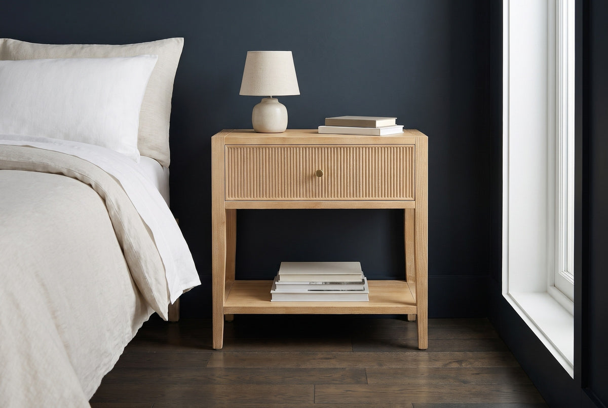 Tonara natural reeded nightstand with a ceramic lamp against a deep blue-charcoal wall in a Brooklyn bedroom