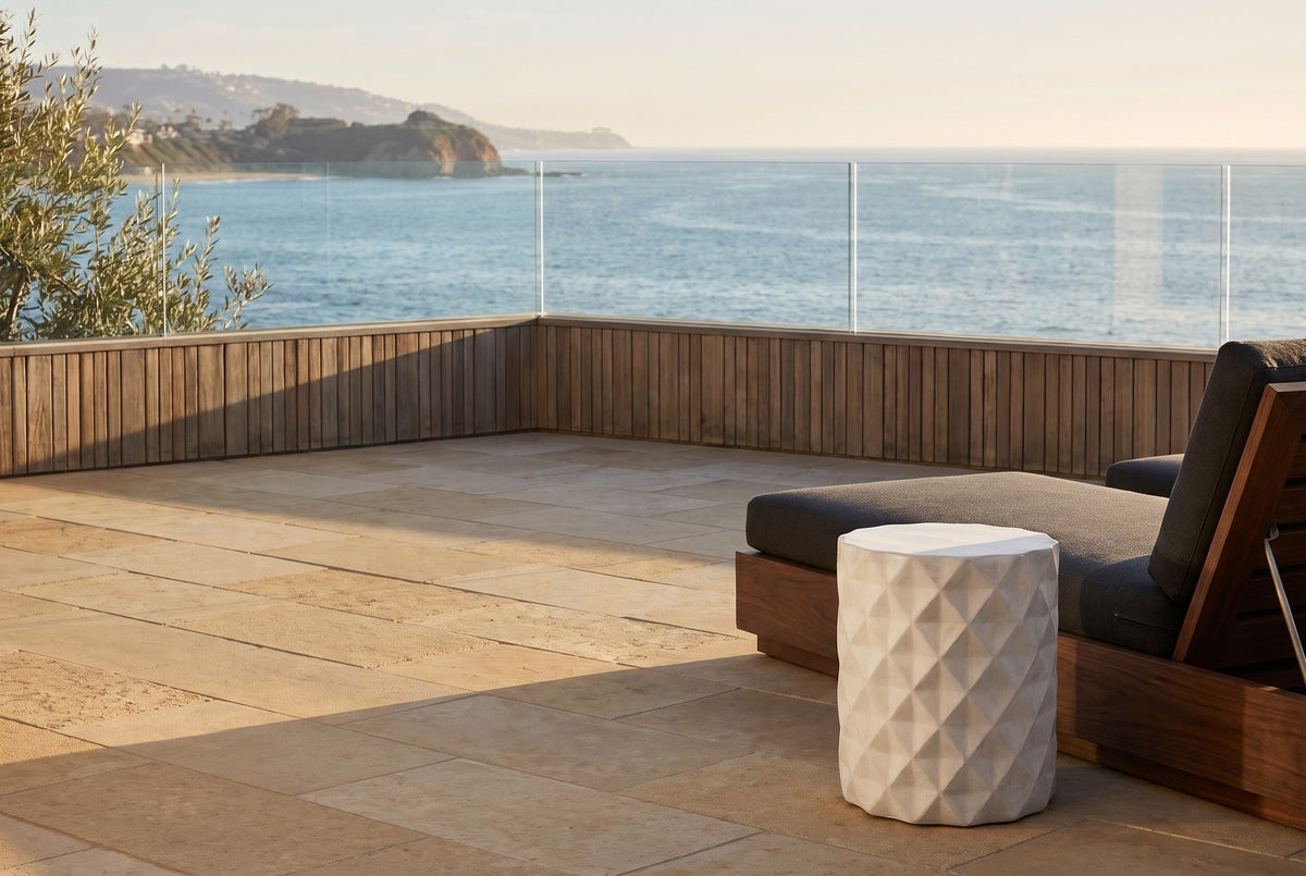 Tortorici antique white faceted concrete drum side table on a limestone terrace in Laguna Beach, a teak slat wall and a glass rail behind, a walnut lounge chair beside it (chair not included)