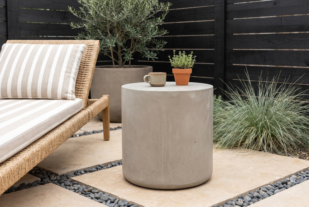 Trabia light grey concrete smooth drum side table with a stoneware cup and a pot of thyme beside a rope lounge chair on limestone slabs in an Austin, Texas courtyard, a black cedar slat fence and an olive tree behind (chair and decor not included)