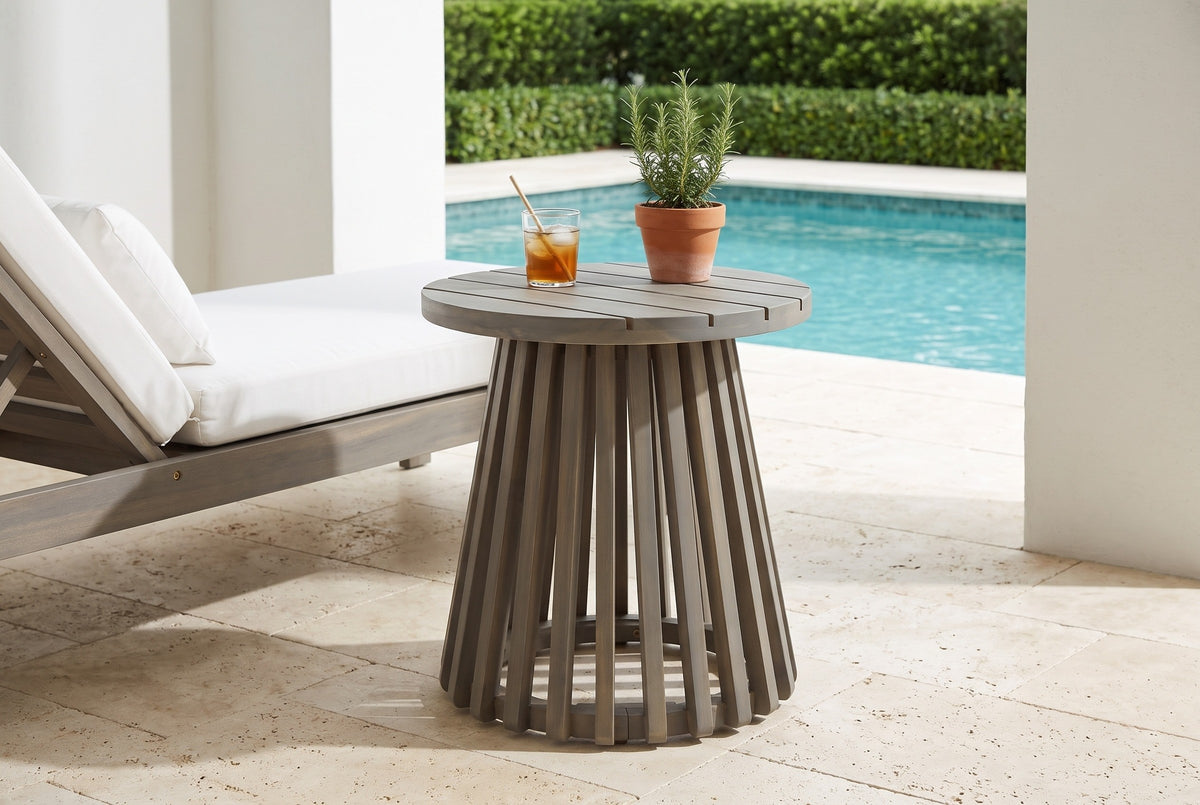 Valderice slatted acacia side table on travertine pavers beside a pool in Naples, Florida, white stucco columns and a clipped hedge behind, a white-cushioned lounge chair beside it (chair, glass and plant not included)