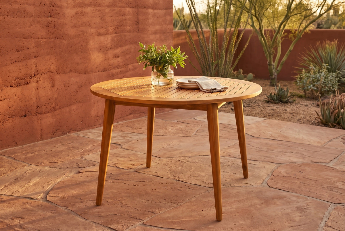 Vittoria round acacia dining table on sandstone flags beside a terracotta rammed-earth wall in Tucson at golden hour, ocotillo and a palo verde behind (vase and plates not included)