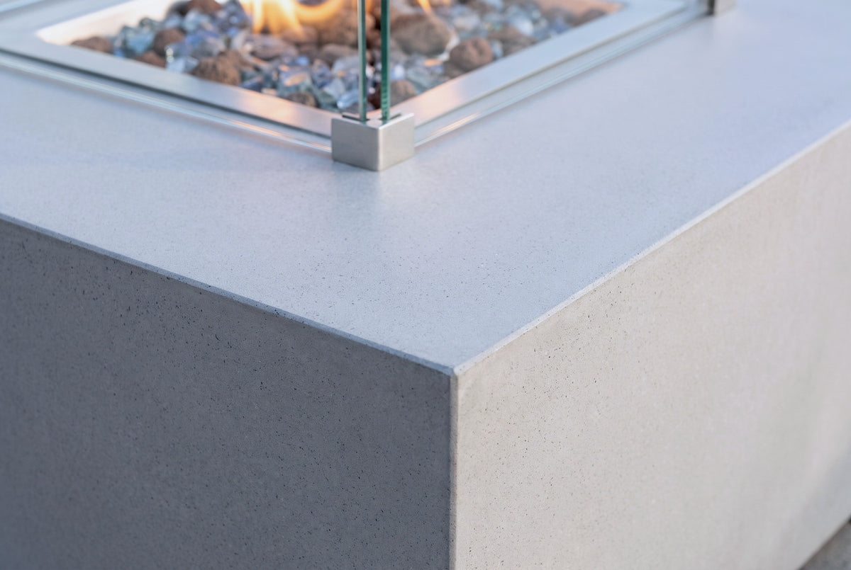 Close-up of the Vulcano fire pit table: the honed light grey concrete top, a square corner and a stainless steel bracket of the glass wind guard above the glass chips and lava rock