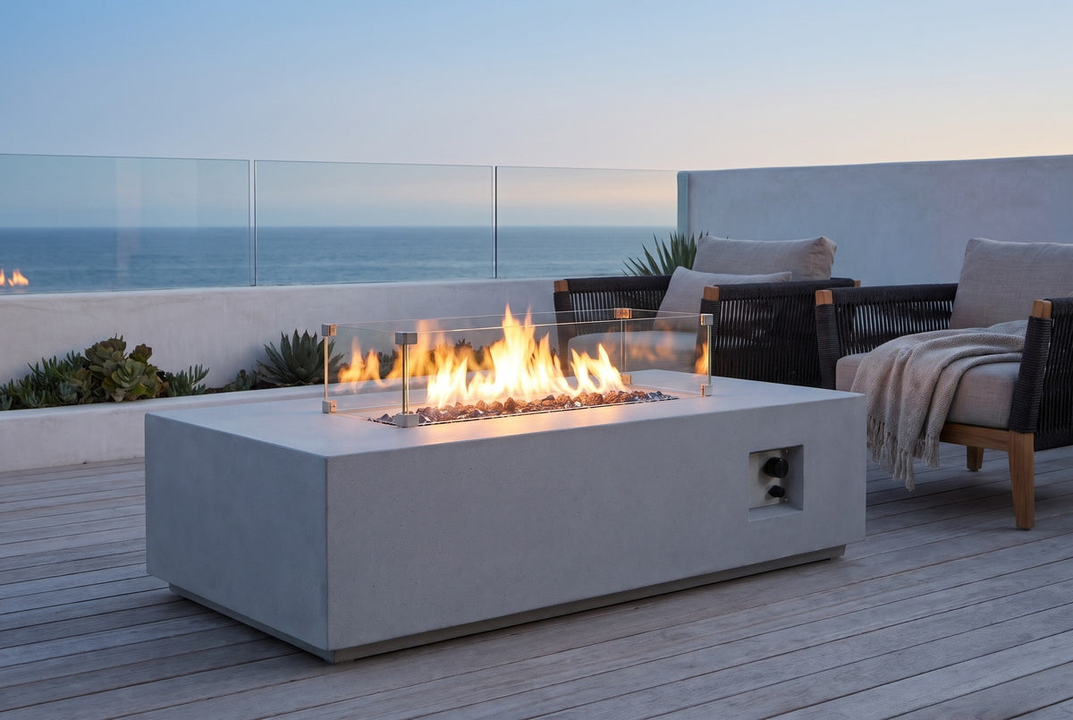 Vulcano concrete fire pit table lit at dusk on a hardwood deck in La Jolla, California, with a glass balustrade, a white wall and the Pacific beyond (chairs and throw not included)