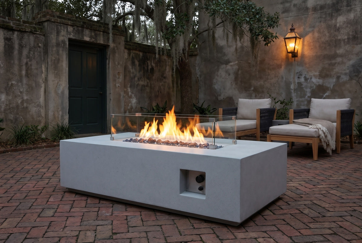 Vulcano concrete fire pit table lit at dusk on a herringbone brick courtyard in Savannah, Georgia, with an old mossy stucco wall, a lantern and live oaks behind (chairs not included)