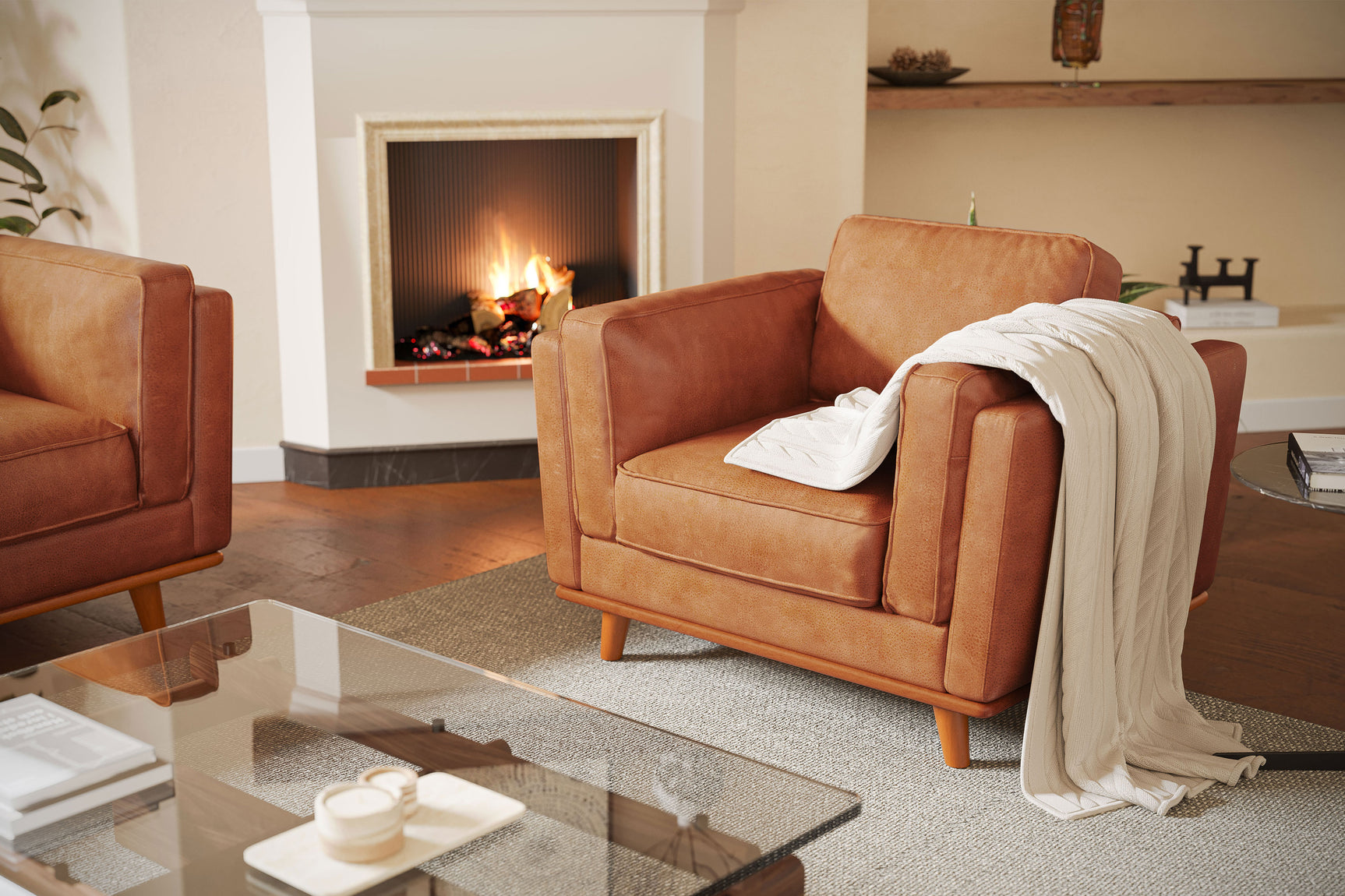 Two tan leather armchairs in a living room setting, one with a white knitted throw draped over it. In the background, there's a lit fireplace. A glass coffee table in the foreground holds books and a small tray with cups.