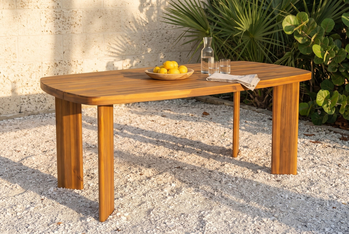 Adrano acacia outdoor dining table on a crushed white shell ground in a St. Augustine garden at golden hour, a cream coquina stone wall, sabal palms and sea grapes behind (chairs not included)