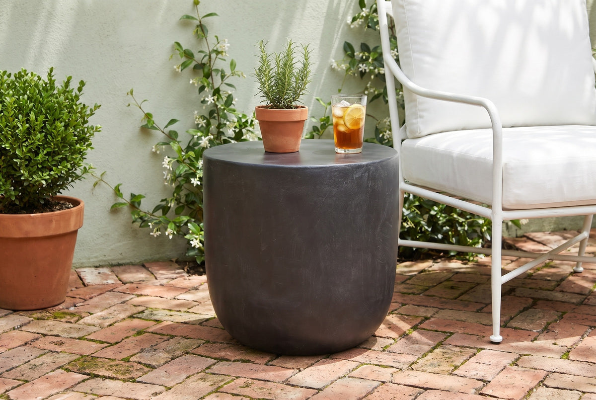 Altavilla matte black side table on herringbone brick in a Savannah garden patio with a sage-green wall and jasmine, beside a white armchair (chair, rosemary and glass not included)