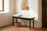 Anagni bench with a folded linen throw against a white adobe plaster wall with an arched niche in a Santa Fe entry hall, with a terracotta tile floor and a seagrass basket