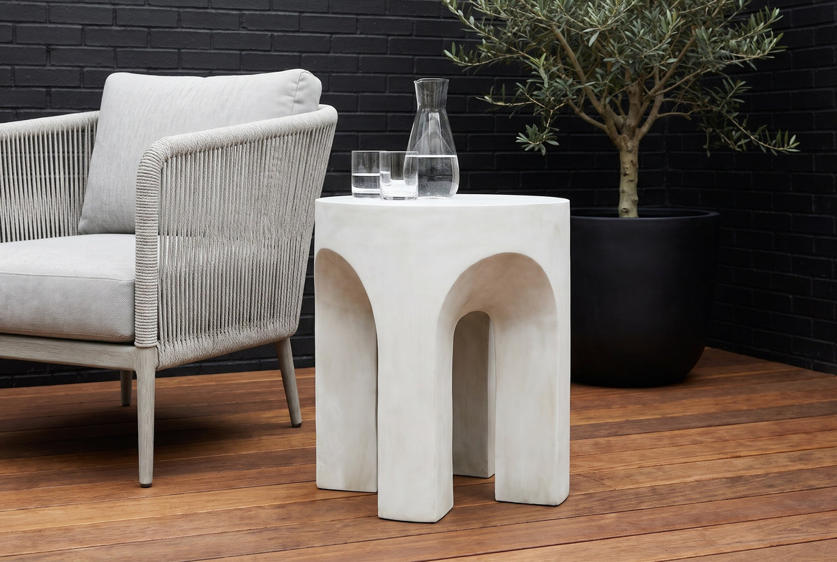 Aragona arched concrete side table with a water carafe and glasses on an ipe deck in a Houston courtyard, a black brick wall and an olive tree behind (chair and decor not included)