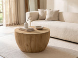 Arona 31.5-Inch Round Solid Fir Storage Drum Coffee Table seen from the side, styled with books, a vase and a stone bowl in front of a cream sofa in a bright plaster room