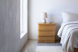 Arzachena oak three-drawer nightstand with a ceramic lamp beside a bed in a Marfa bedroom with a raw concrete wall and a deep-set window