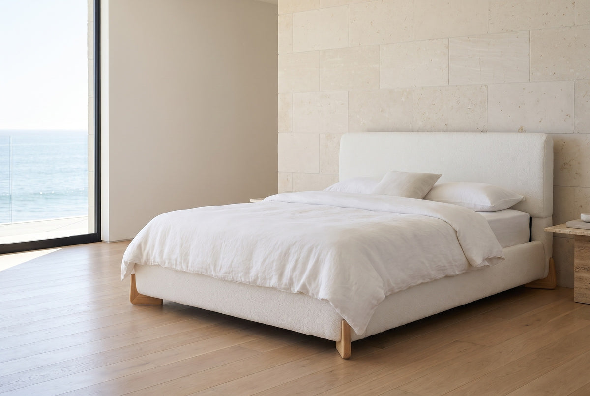 Atessa white boucle bed with wooden corner feet against a pale limestone wall in a Malibu bedroom, a floor-to-ceiling window to the sea at the left