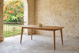 Augusta acacia dining table under a stone loggia with a coral-stone wall and pale limestone floor, a lawn and a flowering tree through the arch (chairs not included, fruit bowl and carafe not included)