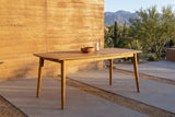 Augusta acacia dining table on concrete pavers on a desert patio at golden hour, a rammed-earth wall, saguaros and mountains behind (chairs not included, bowl and candle holder not included)