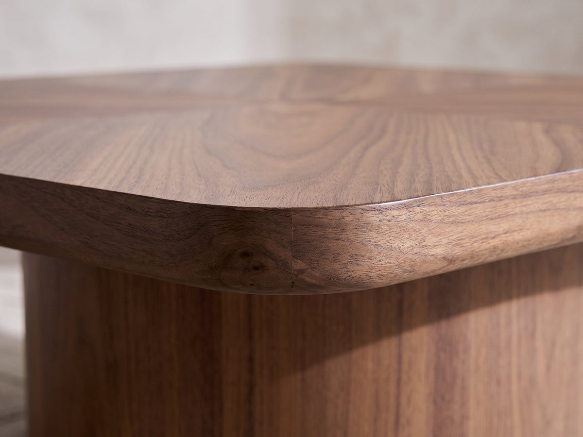 Close-up of the Bari Walnut Veneer Square Pedestal Coffee Table: the rounded corner of the thick walnut top over the drum base