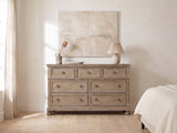 Bassano 65-Inch Wire-Brushed Driftwood Gray 7-Drawer Dresser against a plaster wall under an abstract canvas in a bright bedroom
