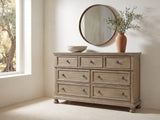 Bassano 65-Inch Wire-Brushed Driftwood Gray 7-Drawer Dresser under a round bronze mirror in a villa bedroom