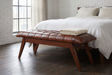 Bevagna cognac leather bench at the foot of a bed with white linen bedding in a New York loft bedroom with a white-painted brick wall and a dark oak floor, a folded cream blanket on the bench