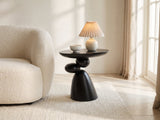 Bitetto Black Fiberglass Stacked Pebble Side Table with a lamp and a ceramic bowl beside a cream boucle armchair in a bright plaster room