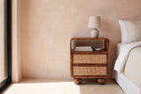 Bitti chestnut mango and cane nightstand with a ceramic lamp beside a bed in an Arizona bedroom with a blush terracotta limewash wall and a tall glass door