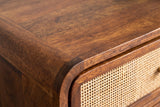 Close-up of the chestnut mango wood on the Bitti nightstand: swirling grain on the rounded top edge beside a cane drawer panel