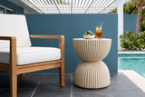 Bivona beige ribbed hourglass side table with a bowl of limes and a glass of iced tea beside a teak lounge chair on a Sarasota, Florida pool terrace, a teal stucco wall, slate pavers and a white pergola (chair and decor not included)