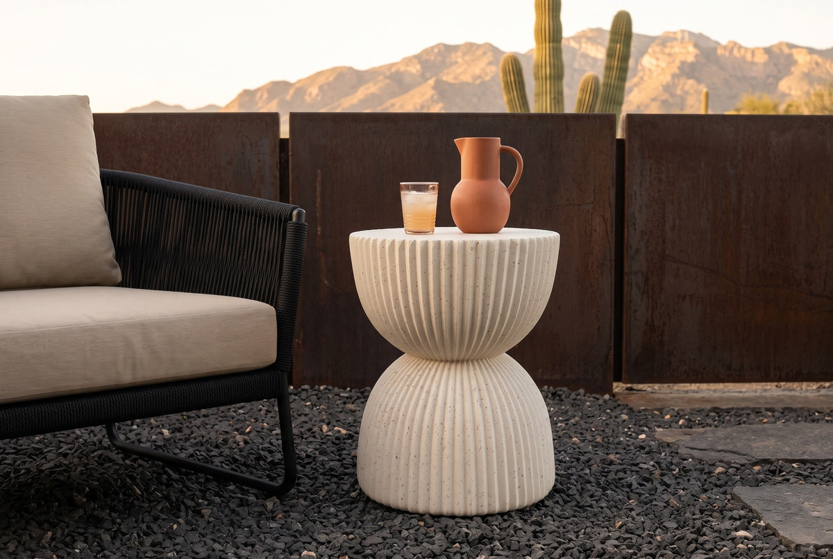 Bivona beige ribbed hourglass side table with a terracotta pitcher and a glass beside a black woven lounge chair on a Tucson, Arizona patio, a rusted steel screen wall, saguaros and mountains behind at golden hour (chair and decor not included)
