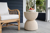 Bivona beige ribbed hourglass side table with a bowl of peaches beside a rattan armchair on a covered porch in Wilmington, North Carolina, a navy shiplap wall, a hanging fern and live oaks beyond (chair and decor not included)