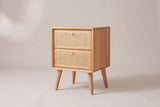 The Bono two-drawer nightstand in light oak with woven cane drawer fronts and gold knobs, three-quarter view on a light studio backdrop