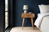 Bonorva light mango one-drawer nightstand with a ceramic lamp beside a bed in a Seattle bedroom with a deep navy wall and a tall black-framed window