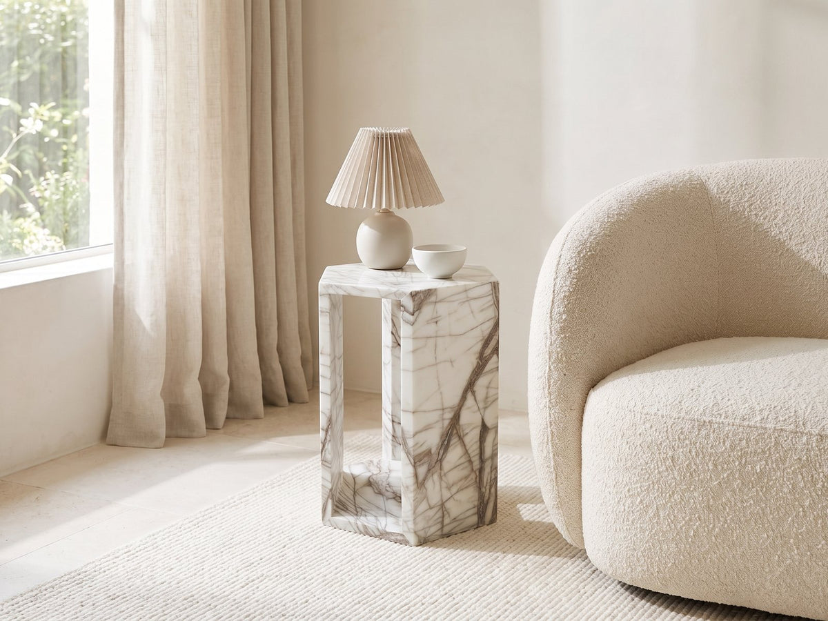 Bovino White Marble Hexagon Side Table with a lamp and a ceramic bowl beside a cream boucle armchair in a bright plaster room
