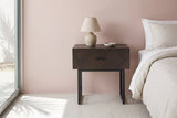 Budoni smoked oak nightstand with a black steel sled base and a ceramic lamp beside a bed in a Palm Springs bedroom with a dusty blush pink plaster wall and terrazzo floor