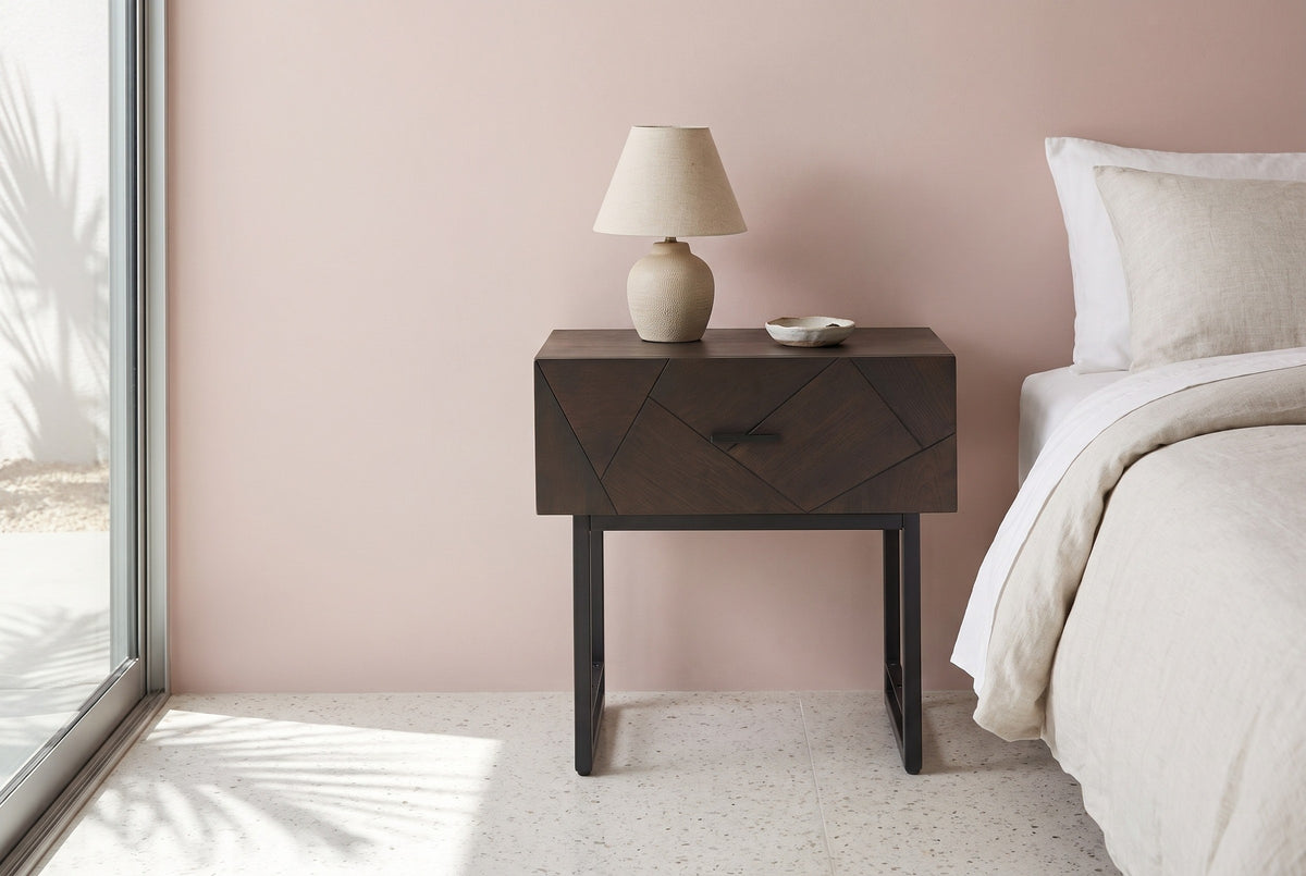 Budoni smoked oak nightstand with a black steel sled base and a ceramic lamp beside a bed in a Palm Springs bedroom with a dusty blush pink plaster wall and terrazzo floor