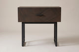 The Budoni nightstand in smoked brown oak, front view on a light studio backdrop: one inlaid drawer with a black bar pull above two black steel sled frames