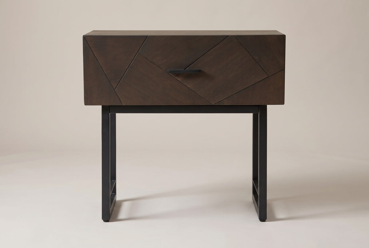 The Budoni nightstand in smoked brown oak, front view on a light studio backdrop: one inlaid drawer with a black bar pull above two black steel sled frames