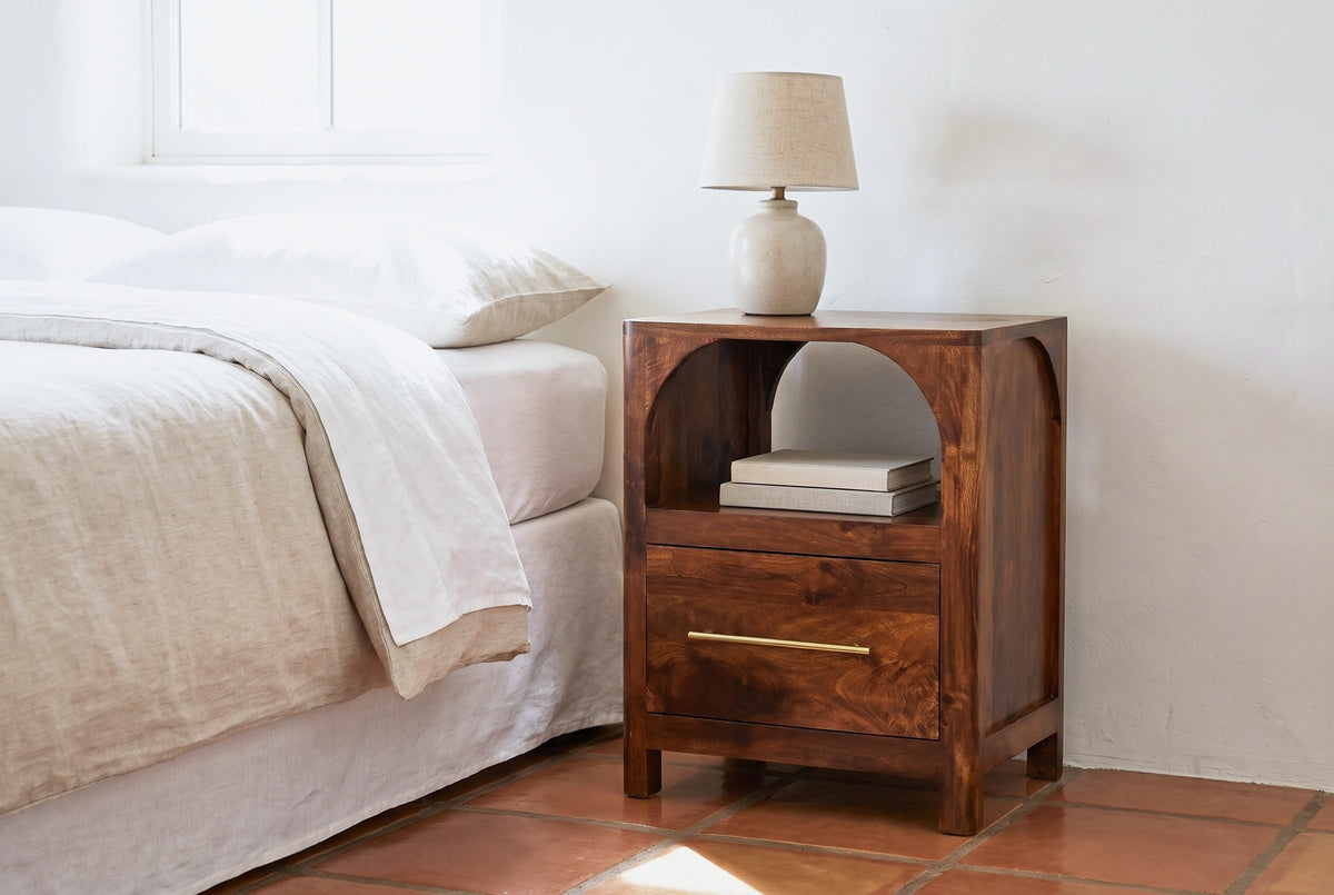 Cabras chestnut mango arch nightstand with a ceramic lamp beside a bed in a Santa Fe bedroom with a white-washed adobe wall and a terracotta tile floor