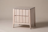 The Calangianus two-drawer nightstand in pale whitewashed hardwood with nine-plank drawer fronts and a grey-washed top, three-quarter view on a warm greige studio backdrop