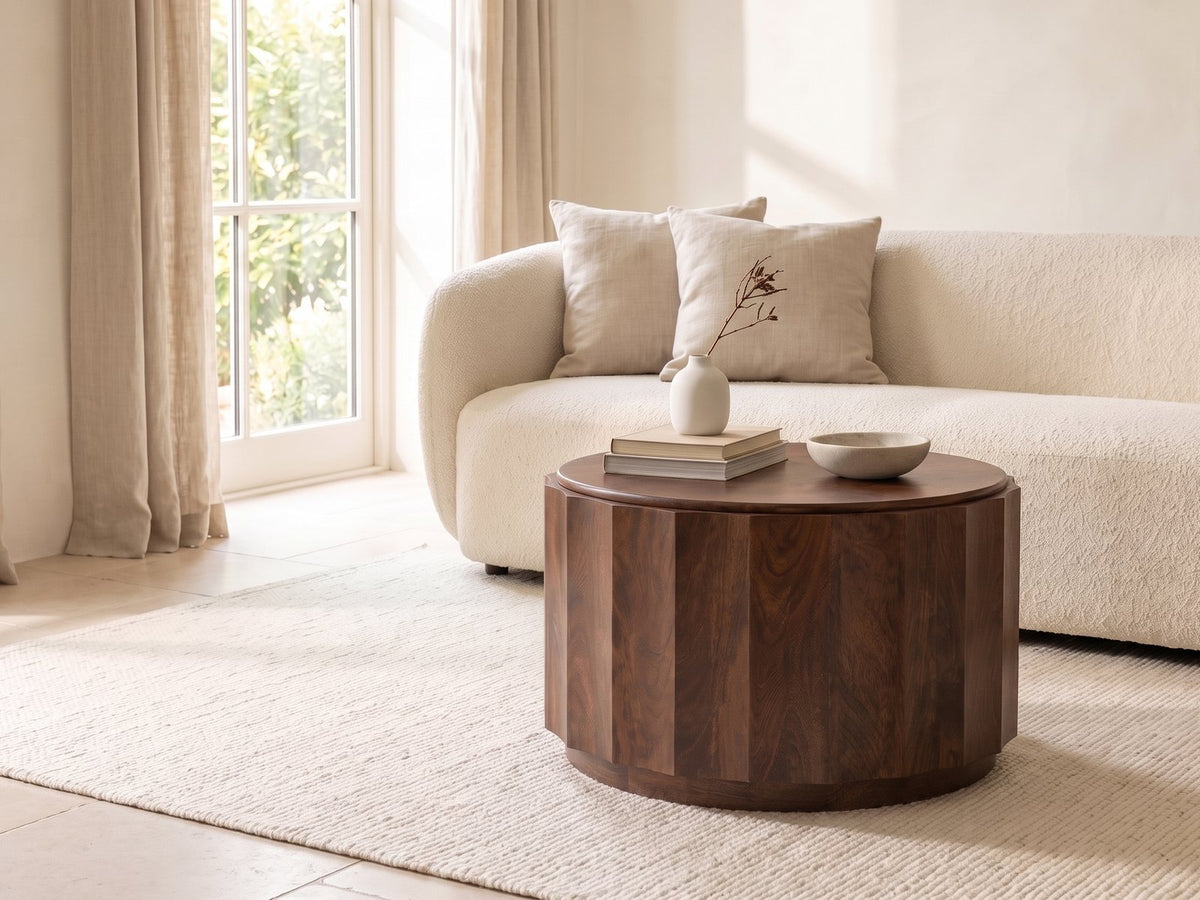 Camaiore Faceted Mango Drum Coffee Table seen from the side, styled with books, a vase and a stone bowl in front of a cream sofa in a bright plaster room