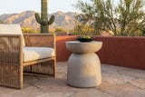 Cammarata light grey concrete dish-top bell side table with a black bowl of limes beside a woven rope armchair on a sandstone patio in Tucson, Arizona at golden hour, a terracotta-red plaster wall, a saguaro and the Santa Catalina mountains behind (chair and decor not included)