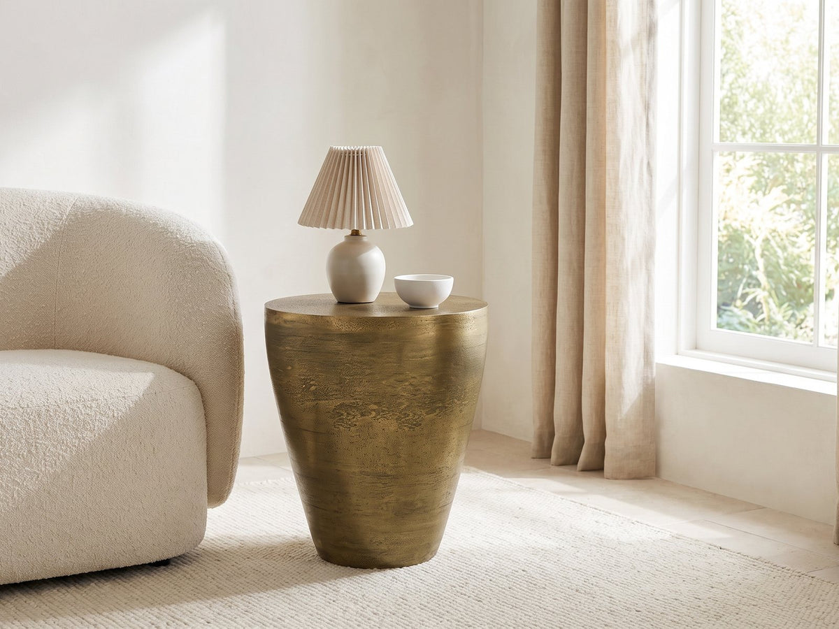 Capestrano Antique Brass Cast Aluminum Drum Side Table with a lamp and a ceramic bowl beside a cream boucle armchair in a bright plaster room