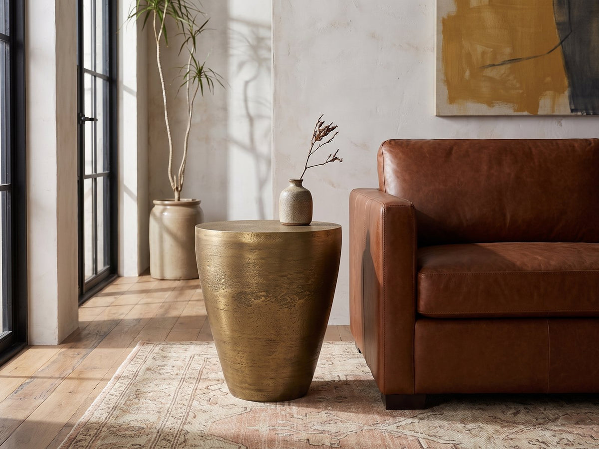 Capestrano Antique Brass Cast Aluminum Drum Side Table beside a leather sofa in an industrial loft