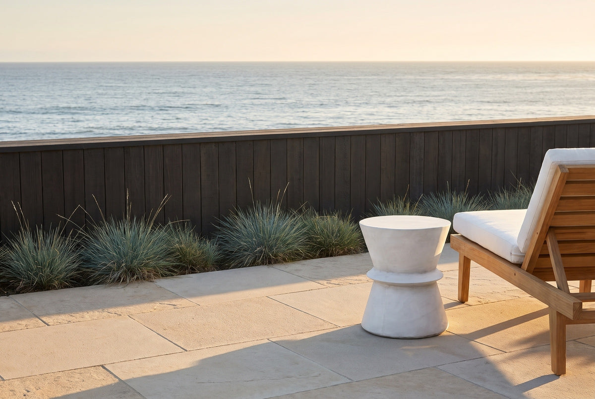 Capizzi antique white concrete hourglass side table on a Laguna Beach terrace above the ocean, a charcoal cedar wall and a teak lounge chair behind (chair not included)