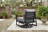 The Carini swivel glider lounge chair seen from behind in a walled Savannah garden courtyard: black aluminum slatted back, teak arm and gray cushions on crushed oyster shell, old brick walls, an iron gate and live oaks with Spanish moss beyond