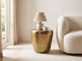 Carovilli Hammered Metal Drum Side Table with a lamp and a ceramic bowl beside a cream boucle armchair in a bright plaster room