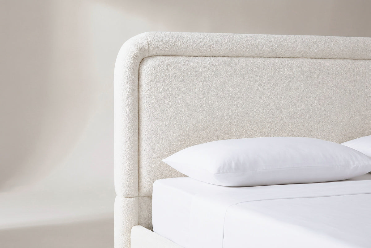 One end of the Casacalenda headboard: a flat white boucle panel inside a rolled border with a rounded top corner, above a white pillow