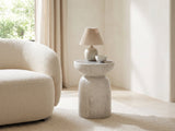 Casarano Fiber Concrete Hourglass Side Table with a lamp and a ceramic bowl beside a cream boucle armchair in a bright plaster room