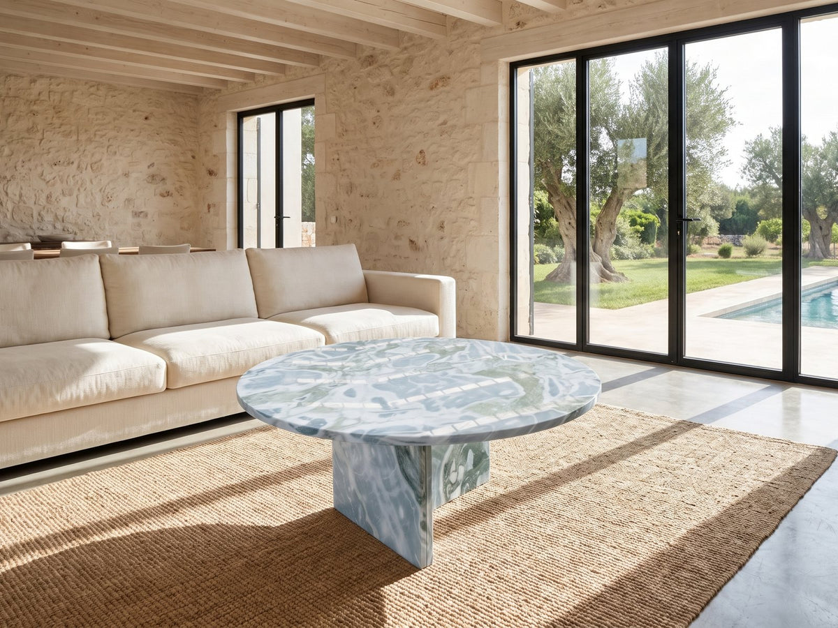 Cascia Compact Blue-Grey Marble Coffee Table seen from the side, on a jute rug in front of a linen sofa in a poolside villa