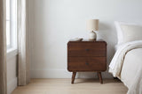 Castelsardo dark walnut two-drawer nightstand with a ceramic lamp beside a bed in a Portland bedroom with a chalk-white limewash plaster wall and a pale oak floor
