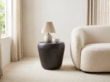 Cervara Rustic Bronze Metal Drum Side Table with a lamp and a ceramic bowl beside a cream boucle armchair in a bright plaster room