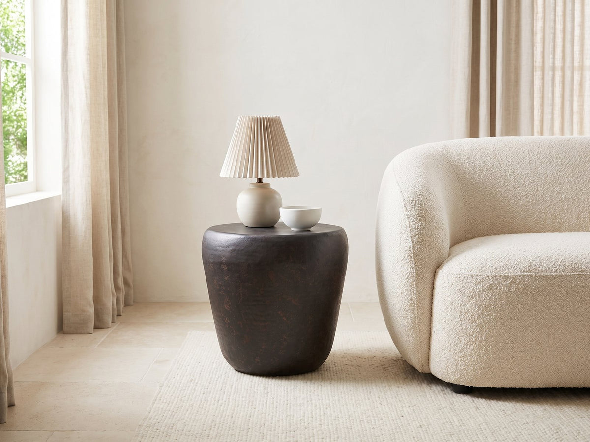 Cervara Rustic Bronze Metal Drum Side Table with a lamp and a ceramic bowl beside a cream boucle armchair in a bright plaster room